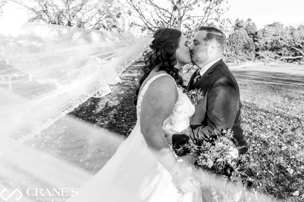 Bride and groom share a romantic kiss at the Morton Arboretum as the bride's veil floats gracefully in the air around them, creating a dreamy, flowing effect. The couple embraces amidst the natural beauty of the Arboretum, capturing a magical wedding moment filled with love and movement.