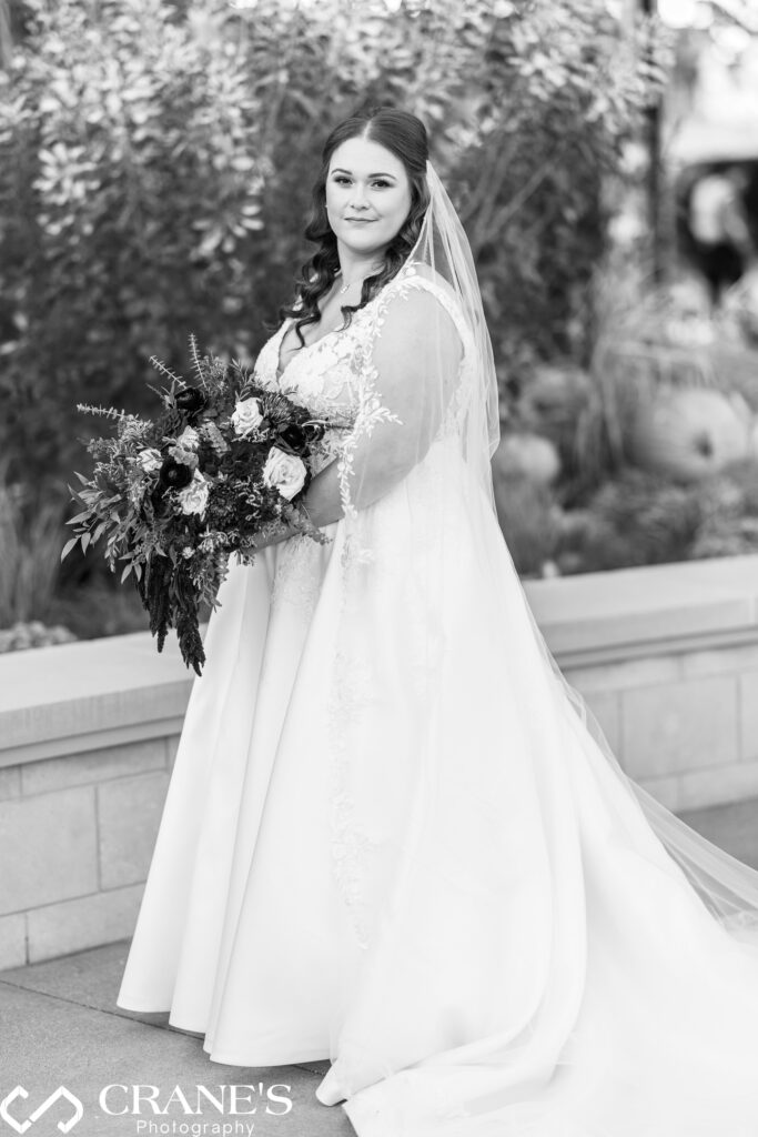 Timeless black and white portrait of a bride at the Morton Arboretum. The bride stands elegantly, wearing a lace gown and veil, with a serene expression as she gazes off-camera. Soft natural light highlights the intricate details of her dress and creates a classic, graceful atmosphere."