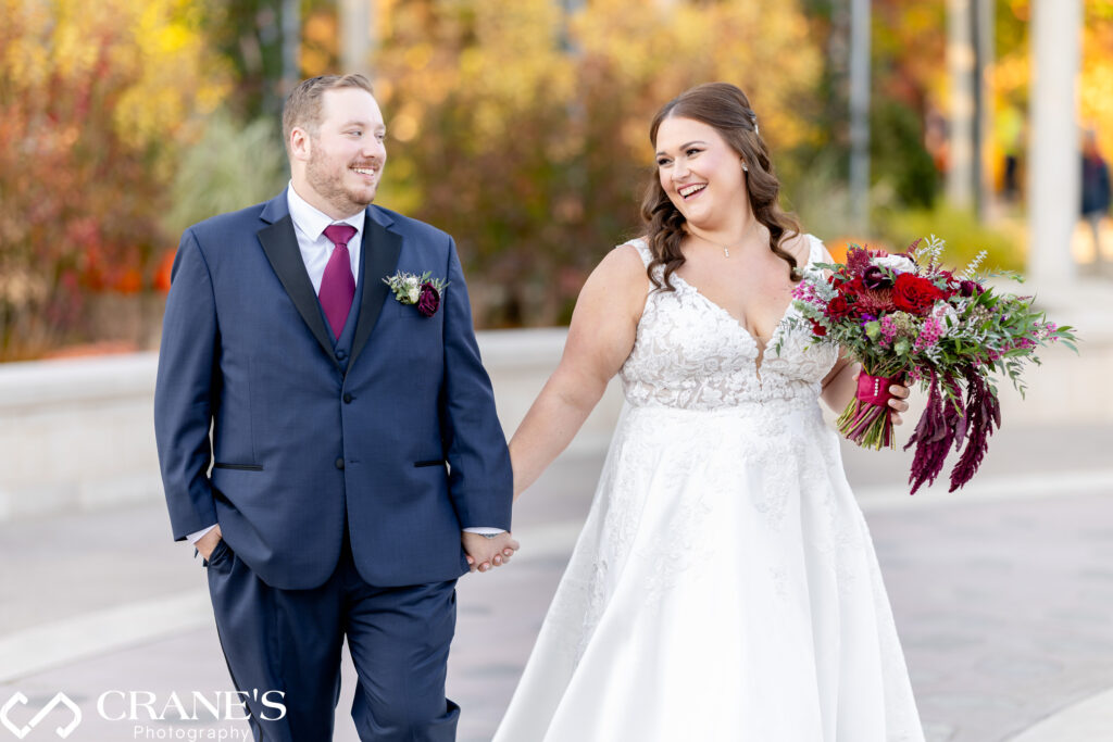 Bride and groom holding hands and smiling at each other as they walk outdoors at the Morton Arboretum. The bride wears a lace gown and carries a bouquet of red and purple flowers with trailing greenery, while the groom, in a navy suit and burgundy tie, looks at her affectionately. The background is filled with soft autumn colors.