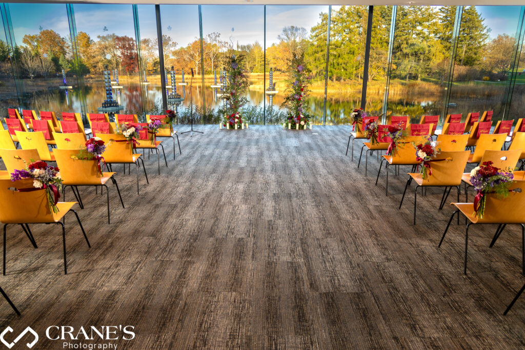 Interior of the Ginkgo Room at Morton Arboretum decorated for a fall wedding ceremony. Warm autumn tones fill the space, with vibrant floral arrangements in shades of burgundy, orange, and gold. Chairs are neatly arranged facing the altar, and sunlight streams through large windows, highlighting the seasonal decor and natural beauty of the surrounding gardens.