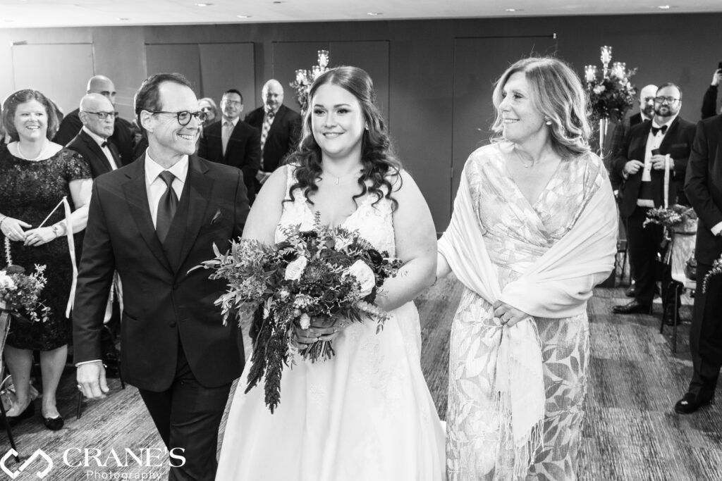 Bride walking down the aisle with her mother and father at her wedding in the Ginkgo Room at the Morton Arboretum. The bride holds her bouquet and beams with joy, while her parents, standing on either side, look at her with proud, smiling expressions. The room is filled with natural light and autumnal decor.