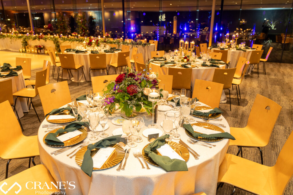 Elegant wedding reception setup in the Ginkgo Room at the Morton Arboretum with fall-themed decor. Tables are adorned with centerpieces in deep reds, oranges, and golds, enhanced by soft lighting. Large windows offer a view of the Illumination Light Exhibit, adding to the enchanting atmosphere of the celebration.