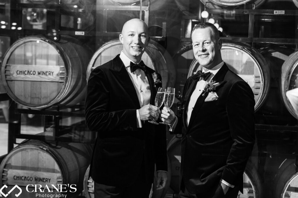 A black-and-white photograph of a same-sex wedding couple dressed in elegant tuxedos, raising champagne glasses in a toast. They are standing in front of stacked wine barrels labeled 'Chicago Winery,' smiling warmly at the camera.