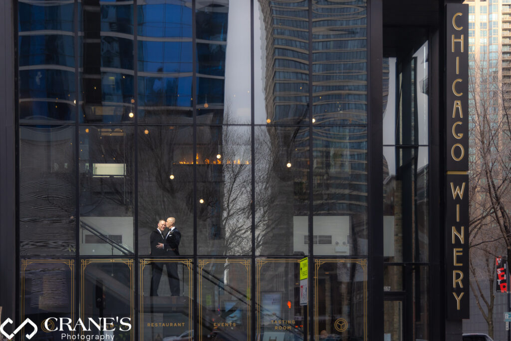 A photograph by Crane's Wedding Photography showing the Chicago Winery building with a couple reflected in the glass facade, the winery's name prominently displayed.