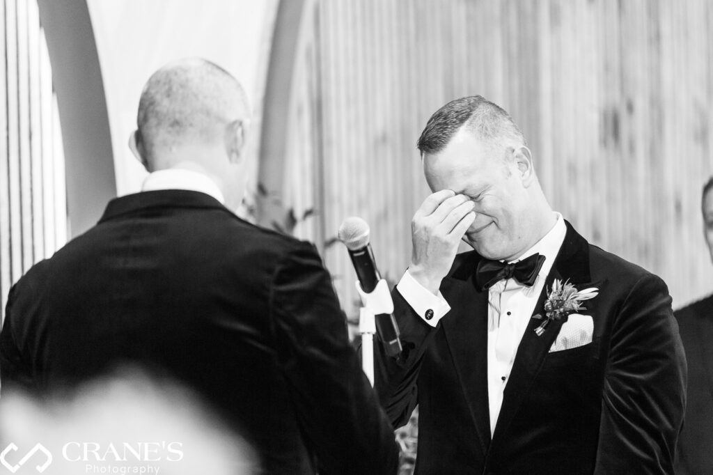 A black-and-white photograph capturing an emotional moment during a same-sex wedding ceremony. One groom, dressed in a black tuxedo with a boutonniere, wipes away tears while his partner, seen from behind, stands at the microphone delivering heartfelt vows.