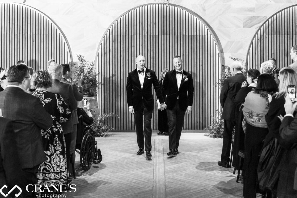 A black-and-white photograph of a newlywed same-sex couple walking hand in hand down the aisle at Chicago Winery after their wedding ceremony. Both grooms, dressed in matching tuxedos, smile as they are cheered on by guests, some capturing the moment on their phones. The venue features elegant arched wooden paneling in the background.