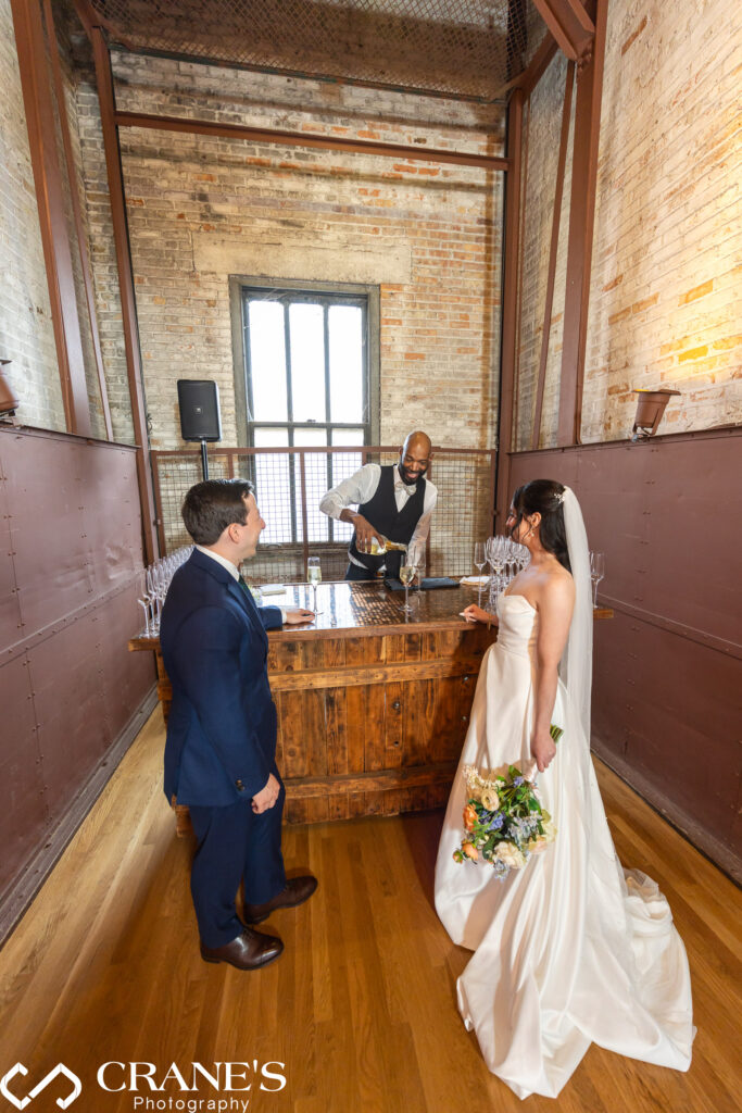 Bride and groom share a joyful moment on their wedding day at the freight elevator bar inside the Skyline Loft at Bridgeport Art Center, as the bartender pours champagne.