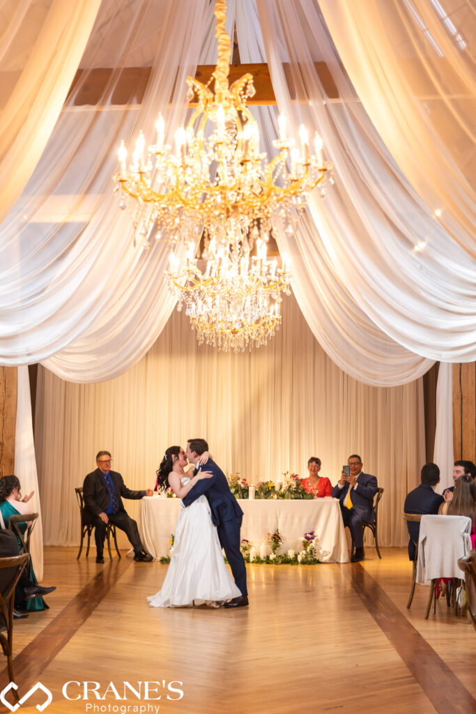 Bride and groom share their first dance on their wedding day under elegant chandeliers and flowing drapery at the Skyline Loft at Bridgeport Art Center, surrounded by family and friends.